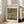 May include: Framed reproduction of Monet's Japanese Footbridge painting featuring soft green and blue tones, showing a curved bridge over a lily pond with water lilies and lush vegetation. The cream-colored exhibition-style poster is displayed in a light wood frame, leaning against a white wall next to a beige upholstered chair in a bright, minimalist living room with large windows and natural lighting.