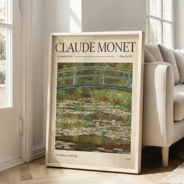 May include: Framed reproduction of Monet's Japanese Footbridge painting featuring soft green and blue tones, showing a curved bridge over a lily pond with water lilies and lush vegetation. The cream-colored exhibition-style poster is displayed in a light wood frame, leaning against a white wall next to a beige upholstered chair in a bright, minimalist living room with large windows and natural lighting.