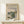 May include: Framed art print featuring Claude Monet's impressionist landscape painting of a river scene with vegetation, buildings, and cloudy sky in soft blues and greens. The beige poster has exhibition-style text layout with a thin wooden frame, displayed on a light wood surface next to a small white wrapped gift with twine and greenery sprig against a cream wall.