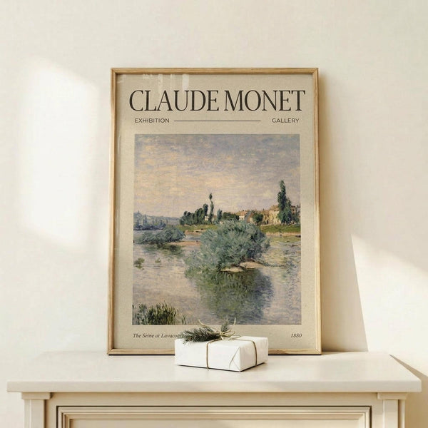 May include: Framed art print featuring Claude Monet's impressionist landscape painting of a river scene with vegetation, buildings, and cloudy sky in soft blues and greens. The beige poster has exhibition-style text layout with a thin wooden frame, displayed on a light wood surface next to a small white wrapped gift with twine and greenery sprig against a cream wall.