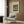 May include: A framed Claude Monet exhibition poster featuring his Japanese Footbridge painting from 1899, displayed above a beige upholstered armchair with geometric patterned pillow. The poster shows the iconic green bridge over lily pads in soft, impressionistic brushstrokes. Room setting includes warm wood paneling, brass floor lamp, and dark round side table with open book.