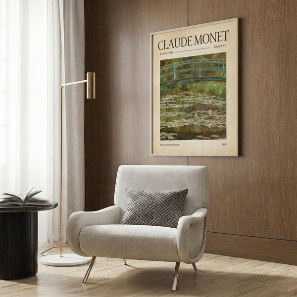 May include: A framed Claude Monet exhibition poster featuring his Japanese Footbridge painting from 1899, displayed above a beige upholstered armchair with geometric patterned pillow. The poster shows the iconic green bridge over lily pads in soft, impressionistic brushstrokes. Room setting includes warm wood paneling, brass floor lamp, and dark round side table with open book.