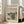 May include: Framed Claude Monet exhibition poster leaning against beige armchair in bright living room. Poster features impressionist river landscape painting with green foliage, water reflections, and soft cloudy sky in muted blues and greens. Cream-colored matting with gallery-style text layout. Natural sunlight streams through tall windows onto light wooden floors.