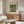 May include: A cream-colored exhibition poster featuring Monet's Japanese Footbridge painting with water lilies, displayed above a walnut wood credenza in a minimalist living room. The poster shows a green bridge over a lily pond in impressionist style. Scene includes a white curved chair, glass oval coffee table, and modern white table lamp on the sideboard.