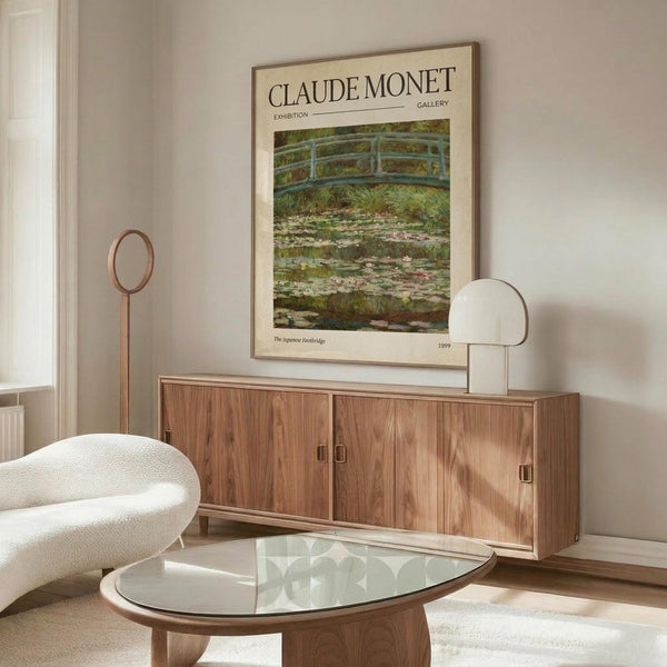 May include: A cream-colored exhibition poster featuring Monet's Japanese Footbridge painting with water lilies, displayed above a walnut wood credenza in a minimalist living room. The poster shows a green bridge over a lily pond in impressionist style. Scene includes a white curved chair, glass oval coffee table, and modern white table lamp on the sideboard.