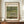 May include: Framed exhibition-style poster of Monet's Japanese bridge painting displayed on wooden console table with white ceramic vase and stacked books. Shows impressionist water lily pond scene with arched footbridge in soft greens and blues. Warm beige wall backdrop with white molding creates gallery-like setting.