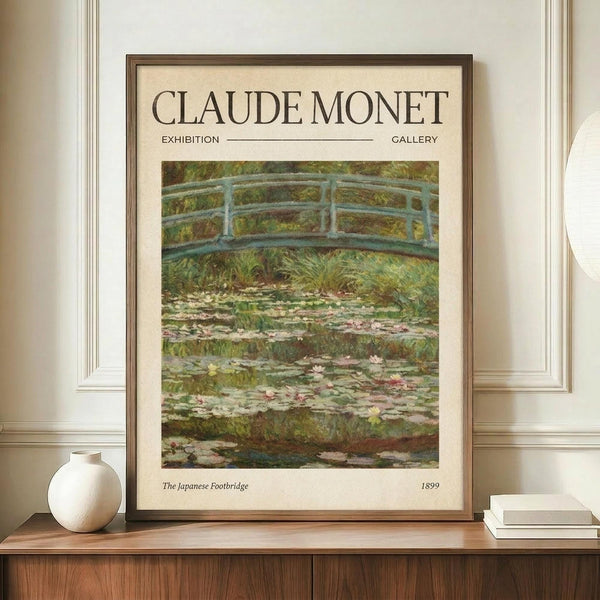 May include: Framed exhibition-style poster of Monet's Japanese bridge painting displayed on wooden console table with white ceramic vase and stacked books. Shows impressionist water lily pond scene with arched footbridge in soft greens and blues. Warm beige wall backdrop with white molding creates gallery-like setting.