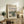 May include: Framed Monet exhibition poster showing an impressionist landscape painting with a winding river, green vegetation, and soft pastel sky. The cream-colored poster is displayed on a rustic wooden sideboard alongside books, a ceramic vase, and a fern plant, with a beige upholstered chair visible in a warm, neutral-toned living room setting.