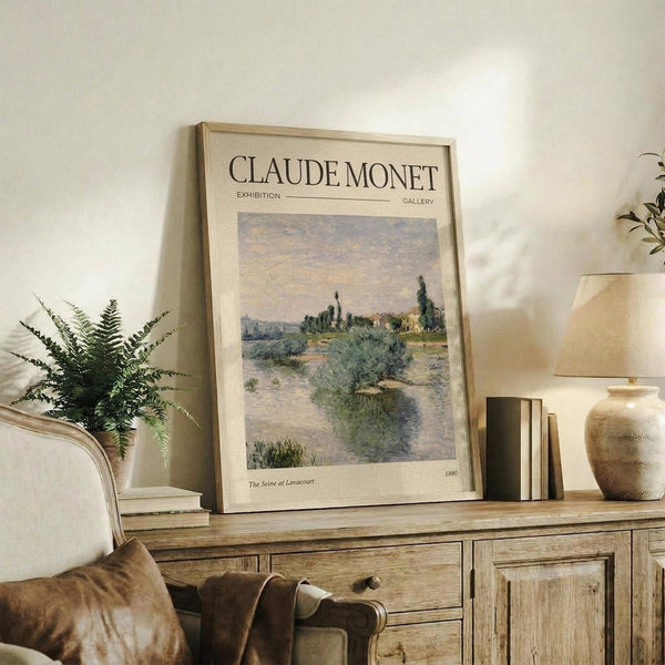 May include: Framed Monet exhibition poster showing an impressionist landscape painting with a winding river, green vegetation, and soft pastel sky. The cream-colored poster is displayed on a rustic wooden sideboard alongside books, a ceramic vase, and a fern plant, with a beige upholstered chair visible in a warm, neutral-toned living room setting.