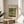 May include: Framed Claude Monet exhibition poster featuring his iconic Japanese bridge painting from 1899, displayed on a light wood round side table next to a beige upholstered chair. The poster shows the famous water lily pond scene with the arched bridge in soft greens and blues. Natural sunlight streams across the neutral-toned room setting with white walls and a plant visible in the corner.
