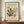 May include: Framed vintage-style exhibition poster featuring abstract botanical cutout shapes in green, orange, and beige tones on cream background, displayed on wooden credenza beside white ceramic vase and books in elegant room with white paneled walls.