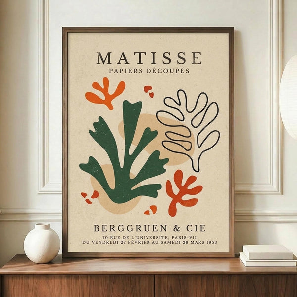 May include: Framed vintage-style exhibition poster featuring abstract botanical cutout shapes in green, orange, and beige tones on cream background, displayed on wooden credenza beside white ceramic vase and books in elegant room with white paneled walls.