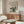 May include: Framed Claude Monet exhibition poster displayed above wooden sideboard in minimalist living room. The vintage-style poster features an impressionist river landscape with soft blues, greens and earth tones. Modern furniture includes cream sculptural chair, glass oval coffee table, and sleek table lamp. Neutral walls and natural lighting create serene gallery-like atmosphere.