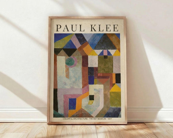 May include: Framed poster print featuring Paul Klee's abstract geometric artwork with colorful triangular shapes at top and architectural forms below in blues, yellows, greens, and earth tones. Natural wood frame against white wall on light wooden floor with soft shadows from window light.