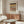 May include: Framed Paul Klee exhibition poster hanging above wood sideboard in modern living room. Abstract artwork features horizontal bands of geometric patterns in earth tones - black, brown, and cream. Room styled with white curved chair, glass oval coffee table, and brass floor lamp. Warm natural lighting creates shadows on white walls.