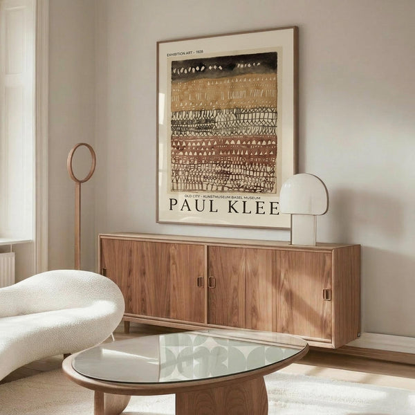 May include: Framed Paul Klee exhibition poster hanging above wood sideboard in modern living room. Abstract artwork features horizontal bands of geometric patterns in earth tones - black, brown, and cream. Room styled with white curved chair, glass oval coffee table, and brass floor lamp. Warm natural lighting creates shadows on white walls.