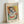 May include: Framed art print in light wood frame leaning against white wall. Cubist-style portrait of a woman with fragmented geometric face in blue, white, and flesh tones. Colorful abstract background with orange, yellow, and green brushstrokes. Text reads "PICASSO" at top with French description below. Frame appears medium-sized against minimalist white background with soft natural lighting creating subtle shadows.