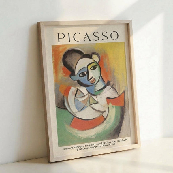 May include: Framed art print in light wood frame leaning against white wall. Cubist-style portrait of a woman with fragmented geometric face in blue, white, and flesh tones. Colorful abstract background with orange, yellow, and green brushstrokes. Text reads "PICASSO" at top with French description below. Frame appears medium-sized against minimalist white background with soft natural lighting creating subtle shadows.