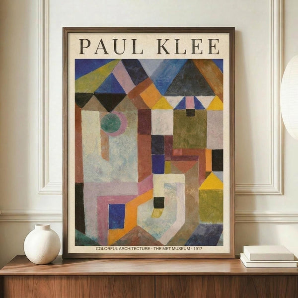 May include: Paul Klee abstract artwork print in brown wooden frame, displayed on walnut sideboard against cream wall with decorative molding. The colorful geometric composition features overlapping houses, triangular roofs, and architectural forms in blues, yellows, oranges, and earth tones. Small white ceramic vase and stacked books sit alongside the framed print.