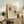 May include: Framed Piet Mondrian abstract art print displayed on rustic wooden sideboard in modern living room setting. The artwork features geometric grid lines with red, blue, and yellow rectangular color blocks on cream background. Light wood frame complements natural decor including fern plant, ceramic vases, stacked books, and neutral upholstered furniture against white wall.