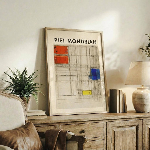 May include: Framed Piet Mondrian abstract art print displayed on rustic wooden sideboard in modern living room setting. The artwork features geometric grid lines with red, blue, and yellow rectangular color blocks on cream background. Light wood frame complements natural decor including fern plant, ceramic vases, stacked books, and neutral upholstered furniture against white wall.