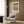 May include: Framed Paul Klee exhibition poster with geometric earth-toned patterns displayed on wood-paneled wall above beige upholstered armchair with geometric pillow. Modern brass floor lamp and dark round side table with open book complete the warm, neutral-toned reading nook setting.