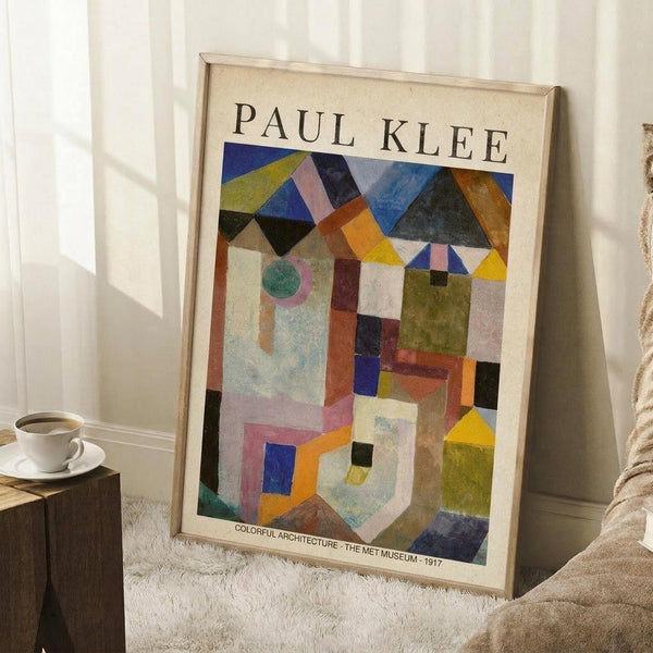 May include: Framed Paul Klee abstract geometric art print leaning against white wall corner. Colorful architectural composition features overlapping triangular roofs, rectangular buildings, and circular elements in blues, oranges, yellows, greens, and earth tones. Light wood frame, positioned on white textured rug next to wooden side table with white coffee cup and saucer. Soft natural lighting creates subtle shadows on white walls.
