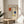 May include: Framed Mondrian-style abstract art print with geometric grid pattern and red, blue, and yellow color blocks on cream background, displayed on round wooden side table next to beige upholstered chair with white throw pillow, green plant leaves visible, soft natural window lighting creating shadows on white wall