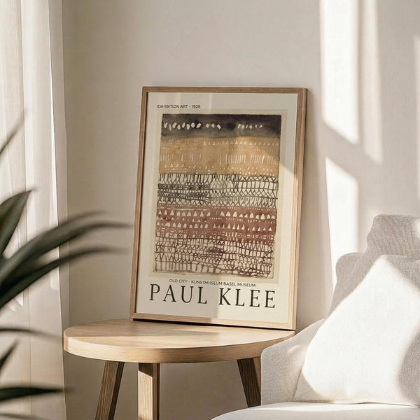 May include: Framed reproduction of Paul Klee's abstract artwork featuring horizontal bands of geometric patterns in earthy brown, beige, and cream tones with triangular and linear motifs. The print displays exhibition text from a 1928 Basel museum show. Shown in thin wooden frame on round wooden side table next to beige upholstered chair with plant leaves visible, all in bright natural lighting against white walls.