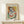 May include: Framed Picasso-style cubist portrait poster featuring abstract female figure with geometric shapes in blues, oranges, yellows and greens, displayed on light wood console table beside small white wrapped gift with twine and greenery sprig against cream wall