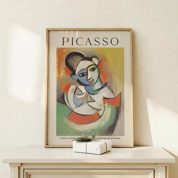May include: Framed Picasso-style cubist portrait poster featuring abstract female figure with geometric shapes in blues, oranges, yellows and greens, displayed on light wood console table beside small white wrapped gift with twine and greenery sprig against cream wall