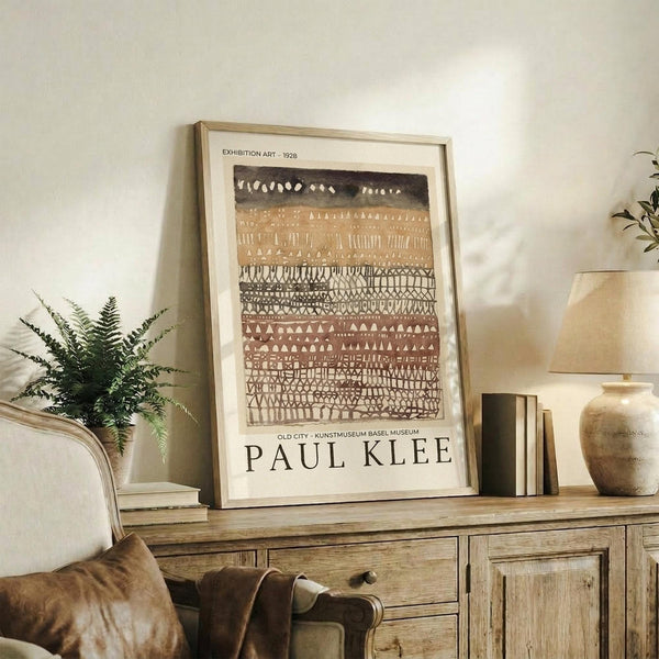 May include: Framed art print with abstract geometric patterns in earth tones - browns, tans, and cream with black accents arranged in horizontal bands. The poster displays in a light wood frame on a rustic wooden sideboard alongside a fern plant, ceramic vase, and books in a warm, naturally-lit room setting.