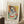 May include: A framed Picasso-style cubist portrait poster featuring a woman with geometric blue, green, and orange color blocks, displayed against a white paneled wall. The artwork shows fragmented facial features in Picasso's characteristic style with bold primary colors. Scene includes a wooden side table with white coffee cup and saucer, white textured rug, and beige upholstered furniture, creating a cozy modern living space with warm natural lighting.