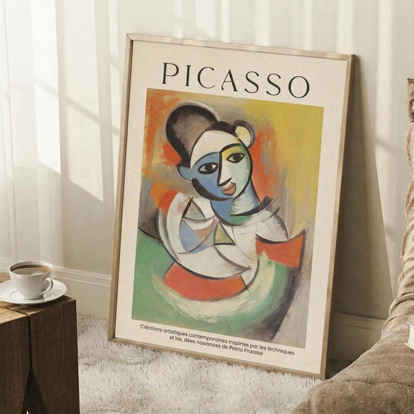 May include: A framed Picasso-style cubist portrait poster featuring a woman with geometric blue, green, and orange color blocks, displayed against a white paneled wall. The artwork shows fragmented facial features in Picasso's characteristic style with bold primary colors. Scene includes a wooden side table with white coffee cup and saucer, white textured rug, and beige upholstered furniture, creating a cozy modern living space with warm natural lighting.