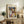 May include: Framed Paul Klee abstract art print displayed on rustic wooden sideboard in neutral-toned living room. Colorful geometric composition features overlapping architectural shapes in blues, oranges, greens, and earth tones. Scene includes fern plant, ceramic vase, stacked books, and beige upholstered chair. Natural lighting creates soft shadows on cream walls.