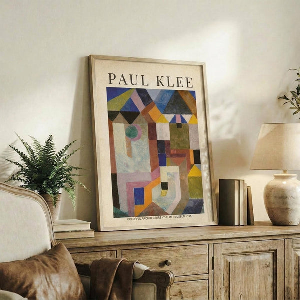 May include: Framed Paul Klee abstract art print displayed on rustic wooden sideboard in neutral-toned living room. Colorful geometric composition features overlapping architectural shapes in blues, oranges, greens, and earth tones. Scene includes fern plant, ceramic vase, stacked books, and beige upholstered chair. Natural lighting creates soft shadows on cream walls.