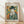 May include: Framed poster print in light wood frame leaning against white wall on wooden floor. Features cubist-style portrait of figure in dark green hat and clothing with white ruffled collar, hand near face in contemplative pose. Geometric background with blue, orange, and yellow squares. Text reads "PICASSO" at top with French description below artwork.