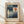 May include: Framed Paul Klee exhibition poster leaning against white wall on light wood floor. Abstract geometric artwork features blue and teal rectangular shapes with orange circle and cream crescent moon against beige background. Thin natural wood frame with cream matting. Soft diagonal shadows cast across white textured wall from natural lighting.