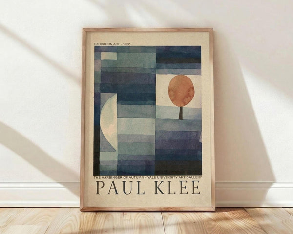 May include: Framed Paul Klee exhibition poster leaning against white wall on light wood floor. Abstract geometric artwork features blue and teal rectangular shapes with orange circle and cream crescent moon against beige background. Thin natural wood frame with cream matting. Soft diagonal shadows cast across white textured wall from natural lighting.