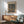 May include: Paul Klee art print in thin wooden frame hanging on white wall above wooden sideboard. Abstract artwork features organic blue and brown shapes resembling stones or cells. Mid-century modern room setting with sage green upholstered chair, white ceramic table lamp, and warm sunlight casting shadows across the scene.