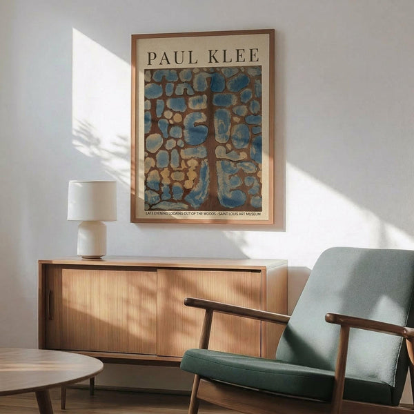 May include: Paul Klee art print in thin wooden frame hanging on white wall above wooden sideboard. Abstract artwork features organic blue and brown shapes resembling stones or cells. Mid-century modern room setting with sage green upholstered chair, white ceramic table lamp, and warm sunlight casting shadows across the scene.