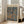 May include: Framed art poster leaning against white wall next to beige upholstered chair in bright living room with large windows. The poster features Paul Klee's abstract painting with blue and brown organic shapes resembling stones or cells against an earthy background. White frame with museum exhibition text at bottom. Natural sunlight creates soft shadows on light wooden floors.