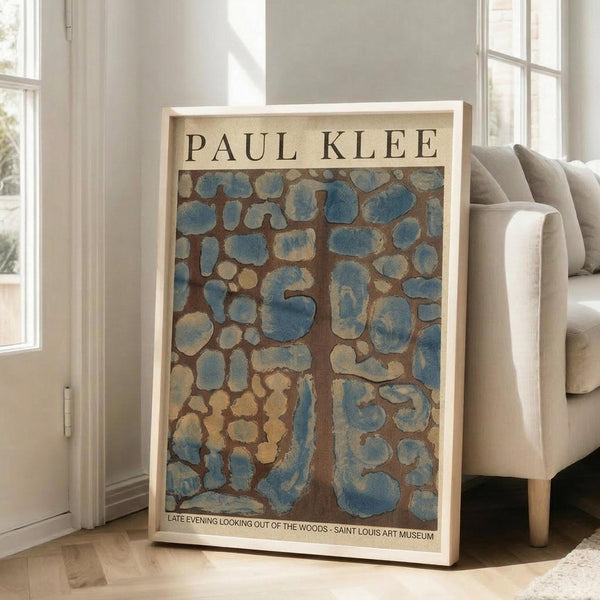 May include: Framed art poster leaning against white wall next to beige upholstered chair in bright living room with large windows. The poster features Paul Klee's abstract painting with blue and brown organic shapes resembling stones or cells against an earthy background. White frame with museum exhibition text at bottom. Natural sunlight creates soft shadows on light wooden floors.