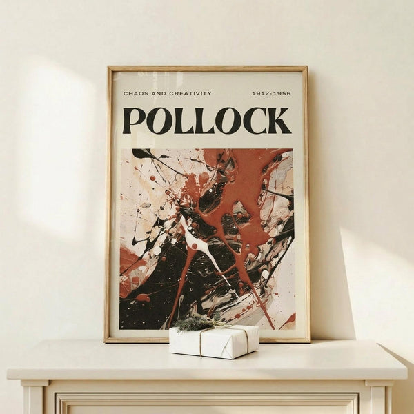 May include: Framed art print featuring abstract expressionist painting with bold black, red, and white paint splatters and drips in chaotic composition. Text reads "CHAOS AND CREATIVITY" and "1912-1956" above "POLLOCK" in large black letters. Natural wood frame sits on light wooden surface beside small white gift box with greenery sprig against cream wall.