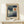 May include: Framed Paul Klee exhibition poster displayed on light wood console table against cream wall. Abstract geometric artwork features blue and teal rectangular blocks with white crescent moon on left, orange-brown circular sun on right. White gift box with green sprig sits in front. Natural wood frame with beige matting. Soft natural lighting creates gentle shadows.