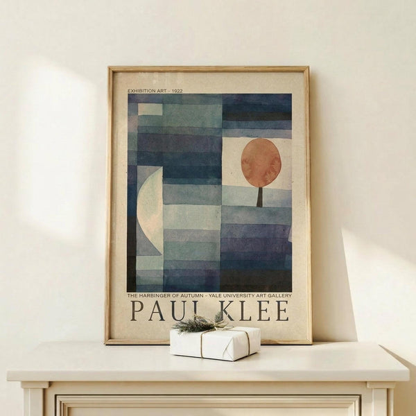 May include: Framed Paul Klee exhibition poster displayed on light wood console table against cream wall. Abstract geometric artwork features blue and teal rectangular blocks with white crescent moon on left, orange-brown circular sun on right. White gift box with green sprig sits in front. Natural wood frame with beige matting. Soft natural lighting creates gentle shadows.