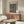 May include: Abstract Paul Klee art print in warm wooden frame hanging above walnut credenza in modern living room. Artwork features organic blue and brown mosaic-like patterns. Room styled with cream curved chair, glass oval coffee table, and white sculptural table lamp. Natural sunlight creates shadows on neutral walls.