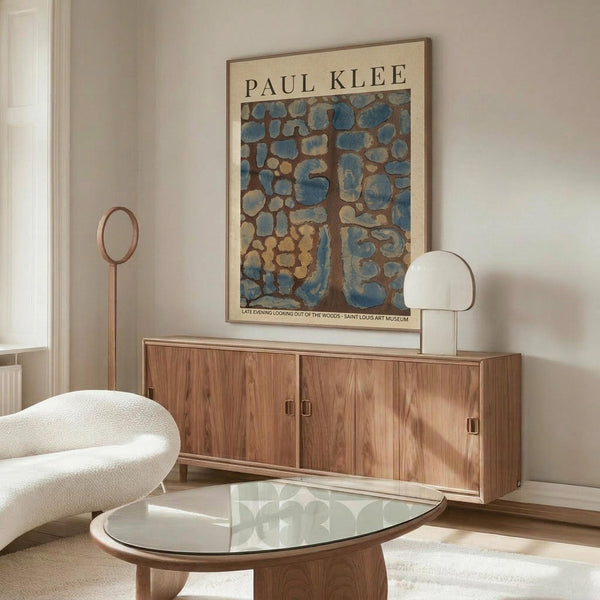 May include: Abstract Paul Klee art print in warm wooden frame hanging above walnut credenza in modern living room. Artwork features organic blue and brown mosaic-like patterns. Room styled with cream curved chair, glass oval coffee table, and white sculptural table lamp. Natural sunlight creates shadows on neutral walls.