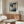 May include: Paul Klee exhibition poster hanging on white wall above wooden sideboard. Abstract artwork features geometric blue and green rectangular shapes with orange circle and white crescent moon. Modern living room setting with cream upholstered chair, glass-top coffee table, and minimalist copper floor lamp. Natural lighting creates warm atmosphere in contemporary interior.