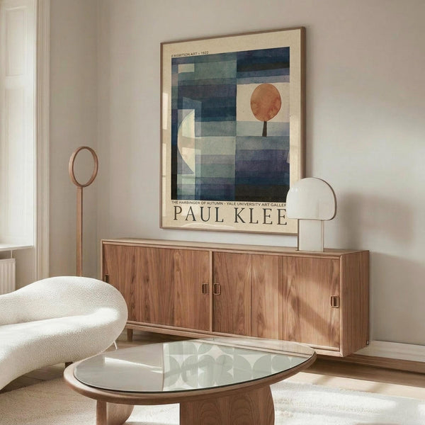 May include: Paul Klee exhibition poster hanging on white wall above wooden sideboard. Abstract artwork features geometric blue and green rectangular shapes with orange circle and white crescent moon. Modern living room setting with cream upholstered chair, glass-top coffee table, and minimalist copper floor lamp. Natural lighting creates warm atmosphere in contemporary interior.